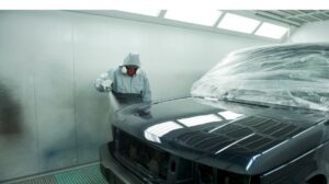 Technician in protective gear spray painting a car in an auto body repair shop.